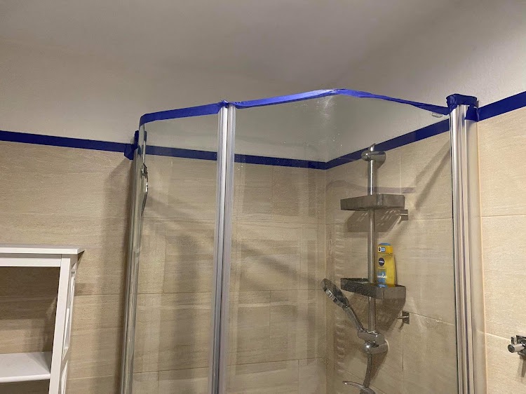 Szklana kabina prysznicowa z niebieską taśmą malarską zabezpieczającą krawędzie przed malowaniem sufitu i ścian w łazience wyłożonej płytkami imitującymi drewno.