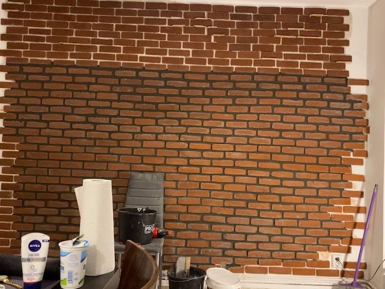 Częściowo wykończona ściana z cegieł klinkierowych, widoczne narzędzia i materiały budowlane, w tym wiadro z klejem i rolka ręczników papierowych.