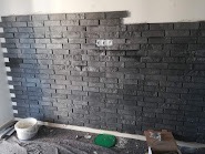 Ściana wyłożona szarą imitacją cegły, widoczne gniazdko elektryczne i resztki zaprawy na podłodze, prace w toku.