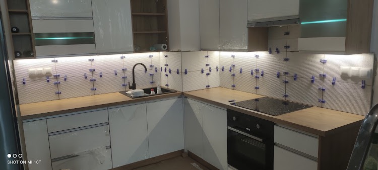 Białe płytki ułożone na ścianie nad blatem kuchennym, z użyciem niebieskich krzyżyków dystansowych, podświetlone listwą LED, z widocznym zlewem i płytą indukcyjną.