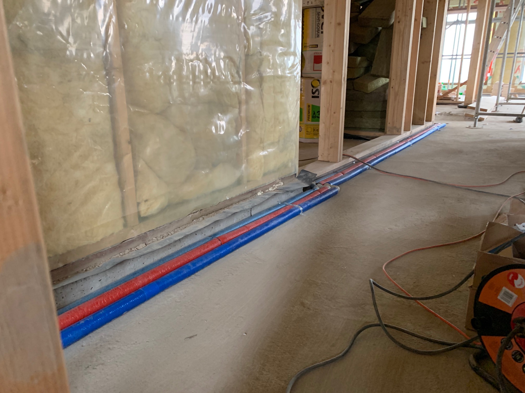 Instalacja rur wodnych i kanalizacyjnych (czerwone i niebieskie) w trakcie budowy domu szkieletowego, widoczne ocieplenie ścian wełną mineralną zabezpieczoną folią.