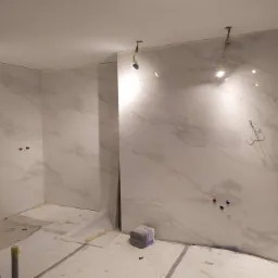 Łazienka w trakcie układania płytek imitujących marmur, widoczne wystające kable oświetleniowe, rury instalacji wodnej i kanalizacyjnej, oraz paczka fugi na podłodze.