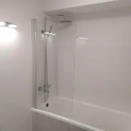 Nowoczesna, jasna łazienka z wanną i szklanym parawanem nawannowym, chromowana bateria prysznicowa z deszczownicą, białe płytki na ścianach i marmurowe wykończenie wanny.