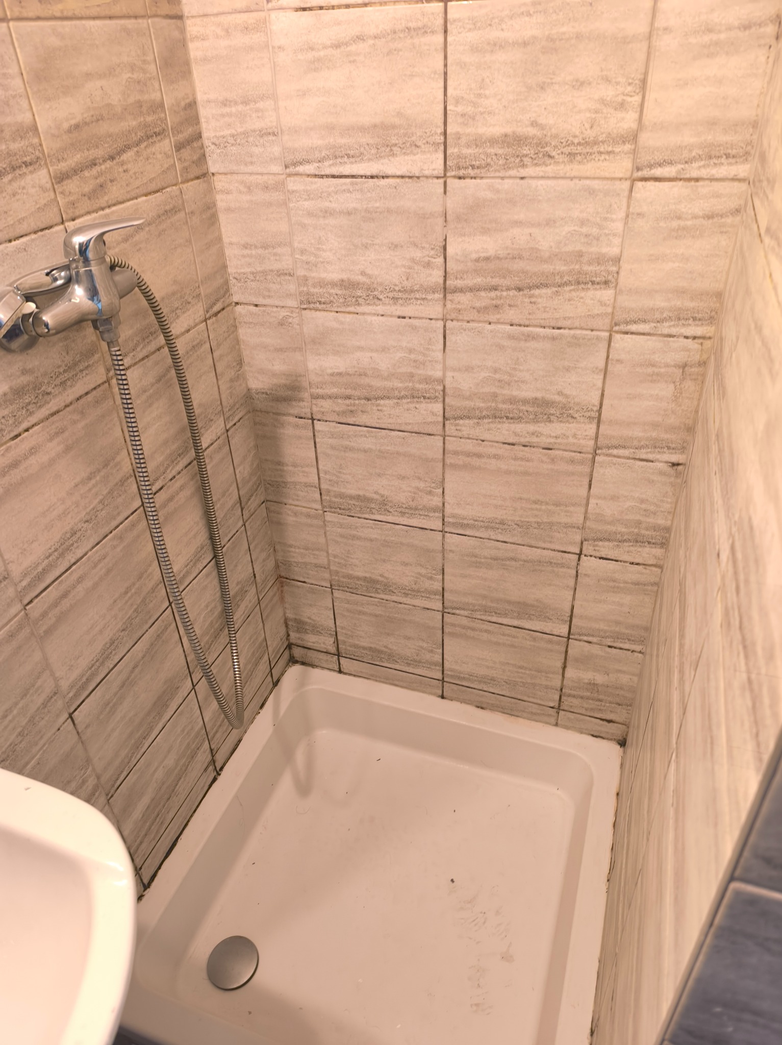 Narożnik kabiny prysznicowej z beżowymi płytkami imitującymi kamień, widoczny chromowany kran z wężem i biały brodzik. Widoczne ślady użytkowania.