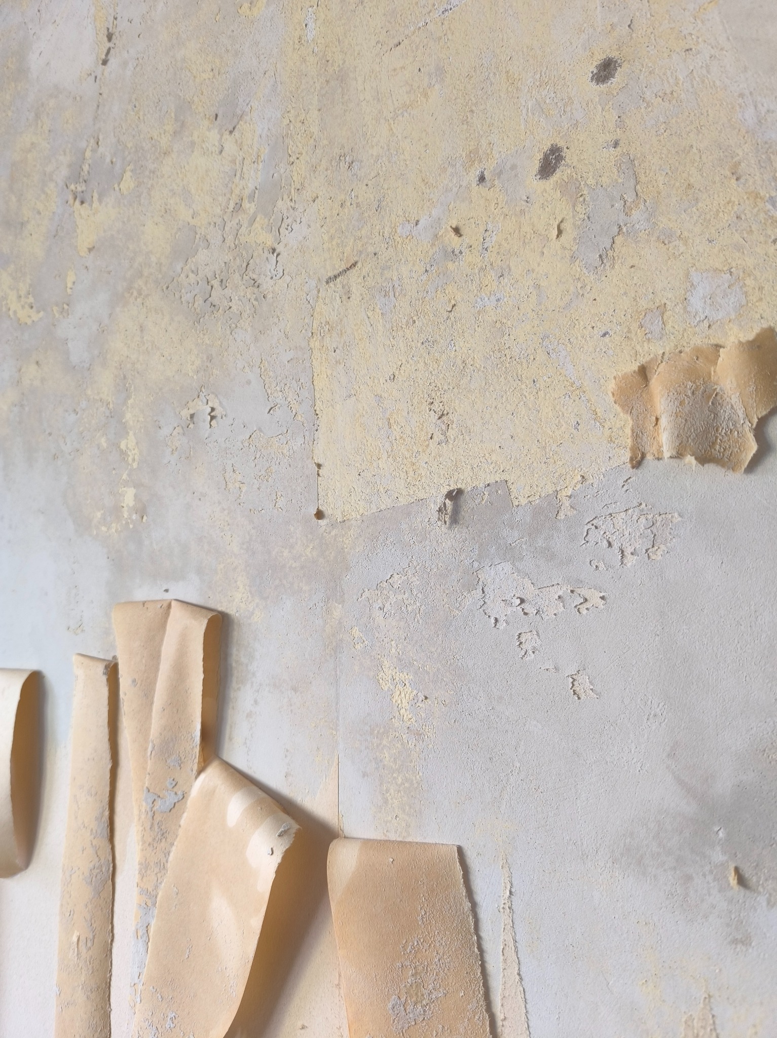 Ściana z resztkami starej tapety i farby, przygotowana do dalszych prac remontowych. Widoczne fragmenty odchodzącej okleiny i nierówności na powierzchni.