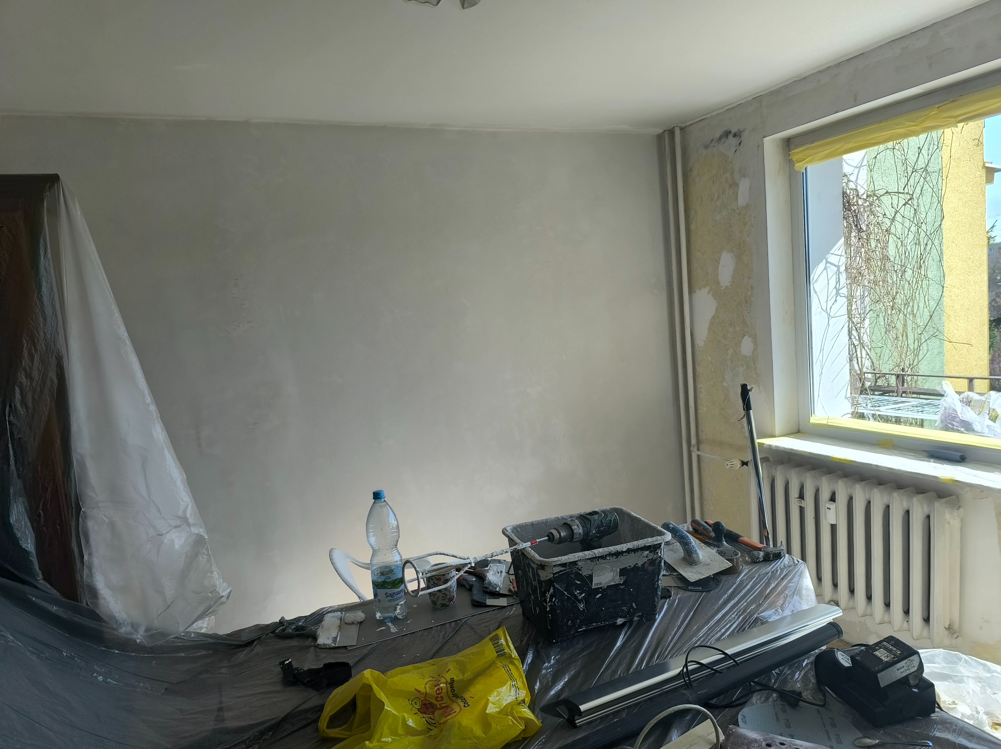 Ściana po nałożeniu gładzi, przygotowana do malowania. Widoczne narzędzia, wiadro z mieszadłem i zabezpieczone folią meble. Okno z roślinnością na zewnątrz.