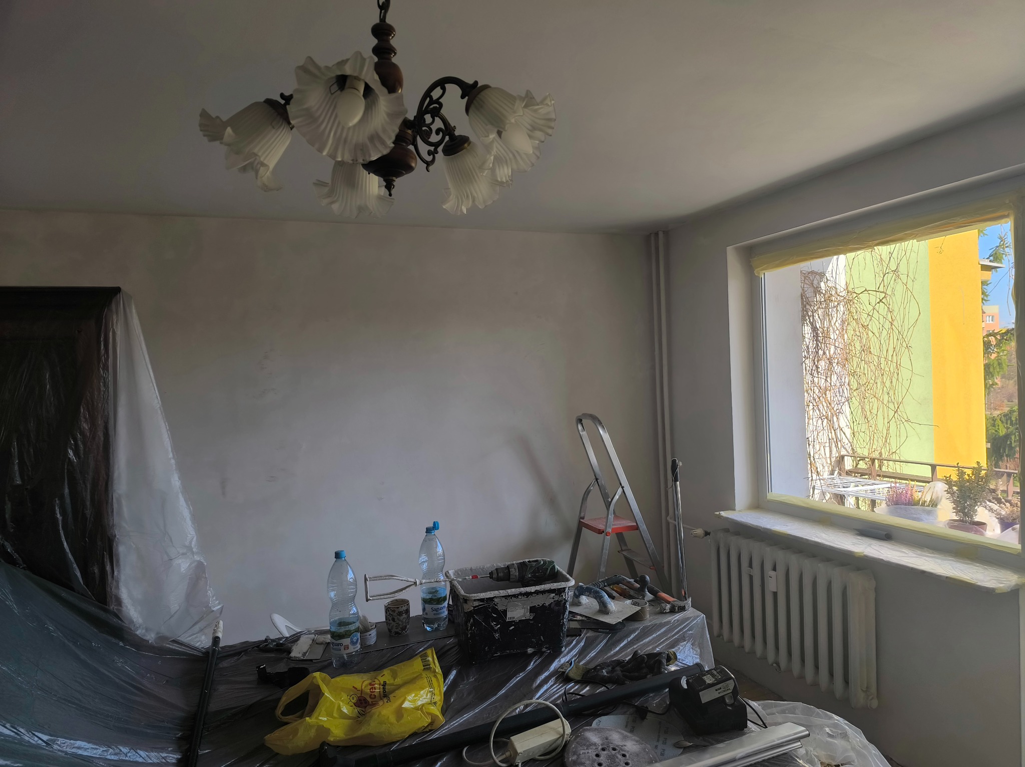 Pokój w trakcie malowania: ściany pokryte świeżą farbą, zabezpieczone meble, drabina i narzędzia malarskie widoczne. Okno z widokiem na zewnątrz.