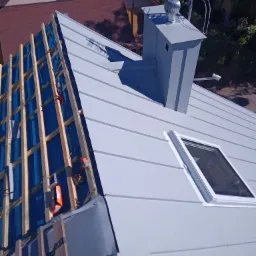 Jasnoszary dach z blachy na etapie montażu, widoczne drewniane elementy konstrukcyjne, okno dachowe, komin z turbiną wiatrową i narzędzia dekarskie na dachu.