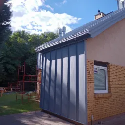 Murowany dom z dobudowaną szarą, metalową konstrukcją, prawdopodobnie w trakcie prac wykończeniowych, widoczne rusztowania i materiały budowlane na trawniku.