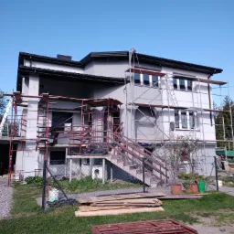 Dwukondygnacyjny dom w trakcie prac elewacyjnych, otoczony rusztowaniami, widoczna drabina oparta o ścianę, na trawniku leżą deski i elementy rusztowania.