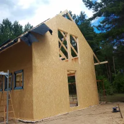 Budowa domu szkieletowego: ściany z płyt OSB, widoczne ramy okienne i fragment dachu z czarną folią. W tle las iglasty i błękitne niebo.
