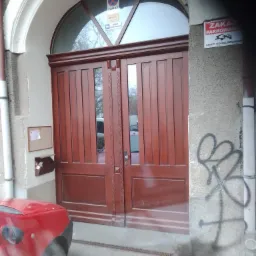 Dwuskrzydłowe, mahoniowe drzwi wejściowe z półokrągłym nadświetlem, osadzone w szarej fasadzie budynku, z widocznym fragmentem czerwonego samochodu po lewej stronie i graffiti na ścianie po prawej.