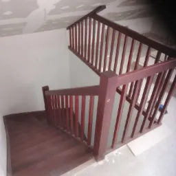 Klatka schodowa z drewnianymi schodami i balustradą w kolorze ciemnej wiśni, widok z g&oacute;ry, sufit w trakcie wykończenia.