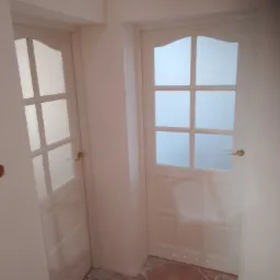 Dwa białe skrzydła drzwiowe z przeszkleniami w stylu angielskim, jedno z klamką w kolorze starego złota, drugie z srebrną, widoczne ślady po remoncie na brązowej podłodze.