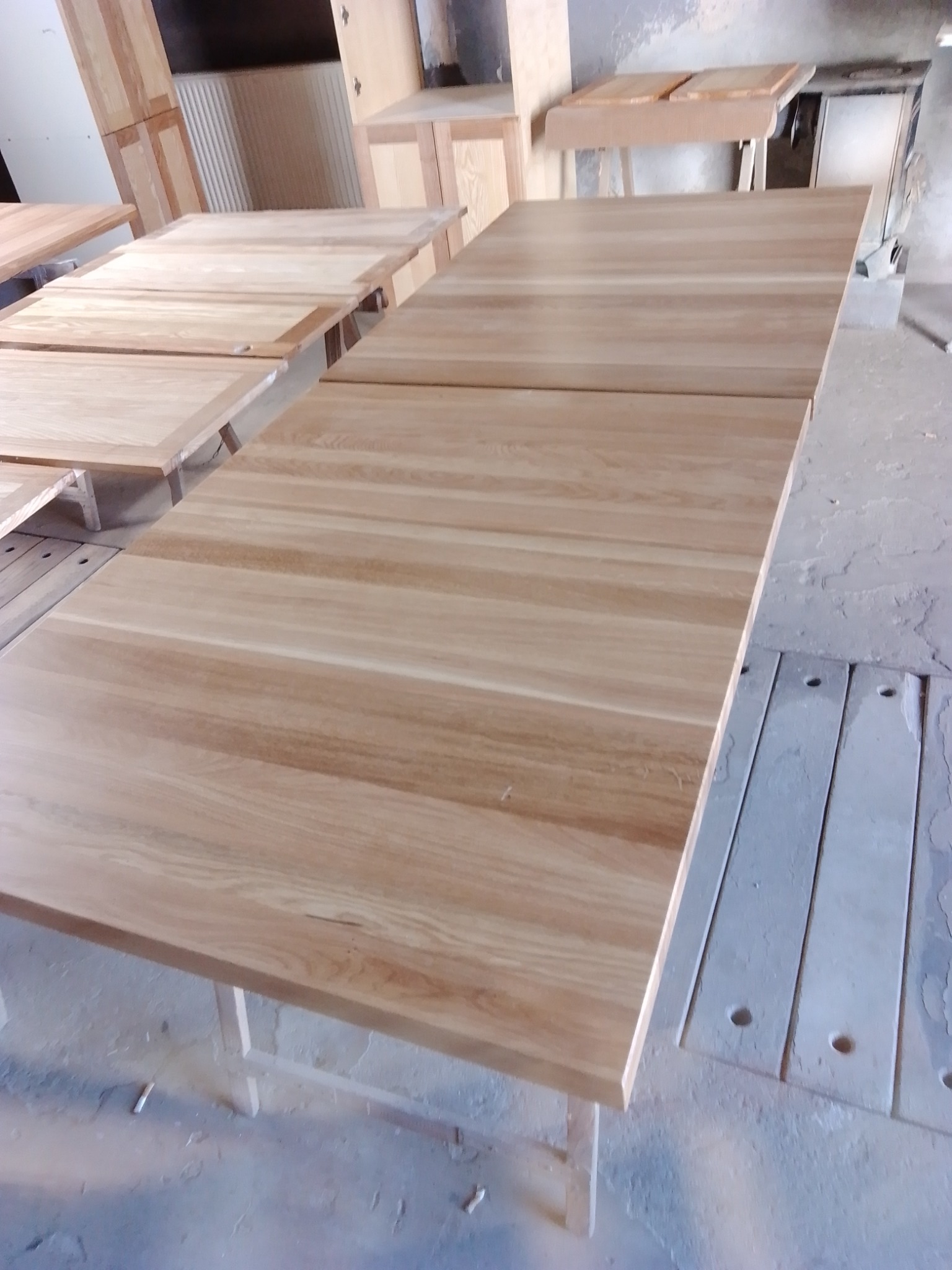 Blaty z jasnego drewna, prawdopodobnie dębowego, ułożone na wspornikach w warsztacie, widoczne słoje i naturalna faktura drewna.