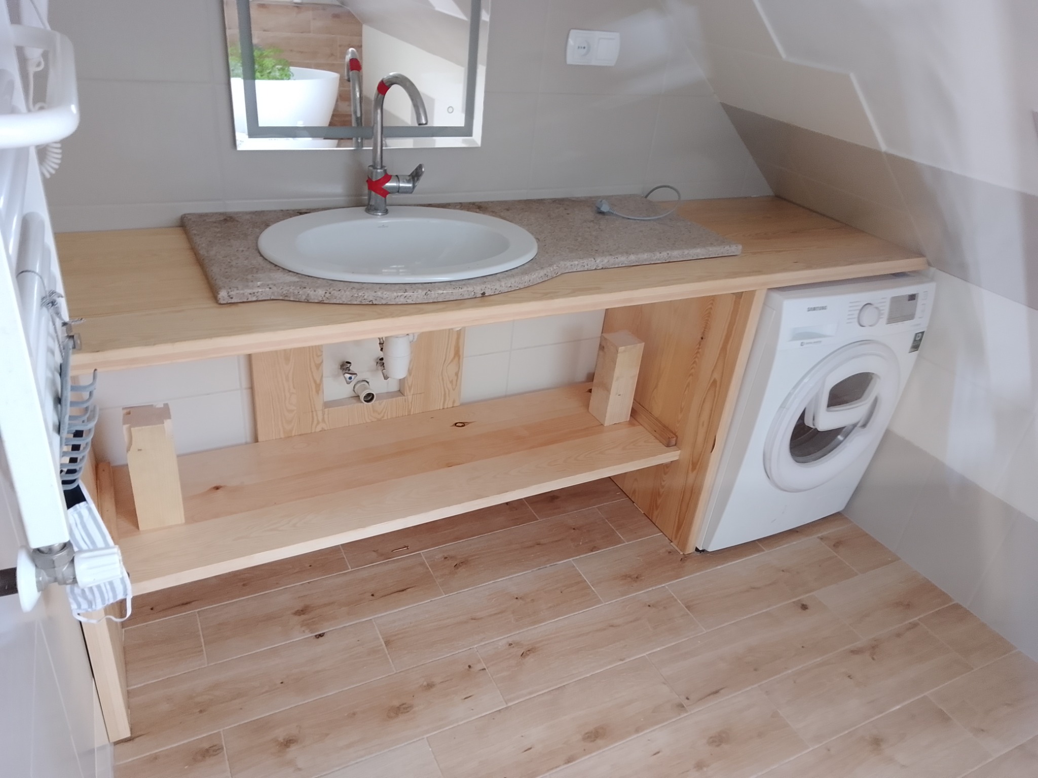 Drewniana zabudowa umywalki z blatem z konglomeratu, z widoczną pralką i lustrem w łazience na poddaszu.
