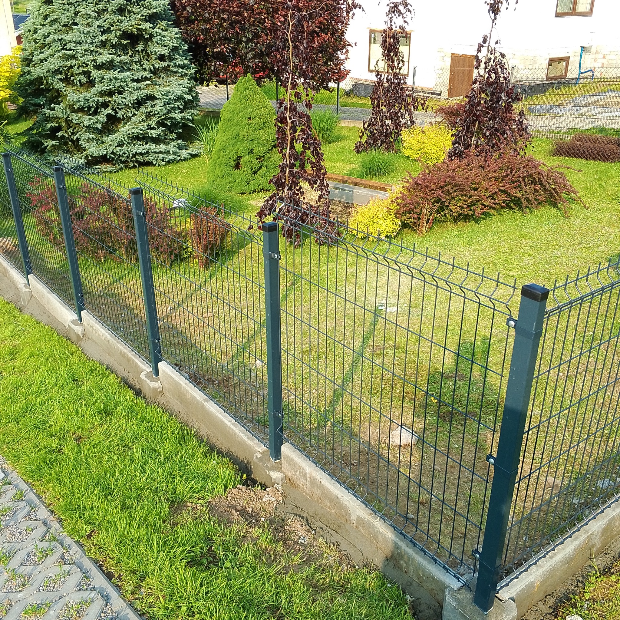 Nowoczesne ogrodzenie panelowe z podmurówką betonową, otoczone zielenią. Widoczne drzewa i krzewy w tle. Świeżo posadzona trawa przy ogrodzeniu.