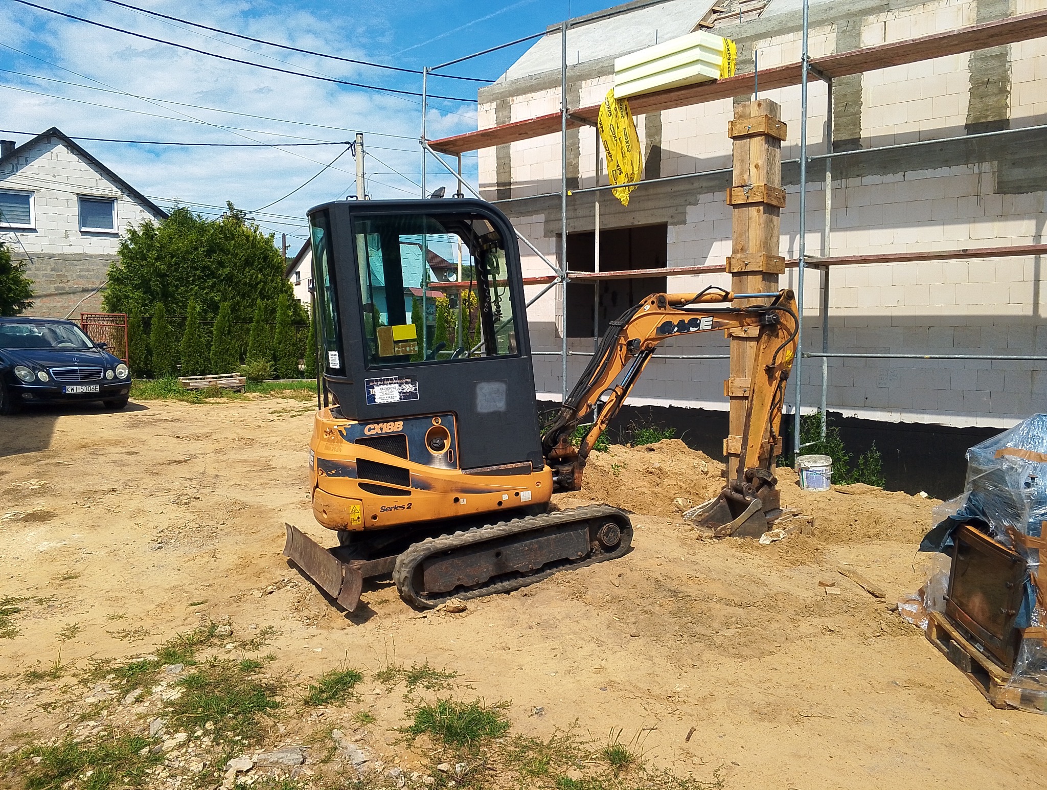 Pomarańczowa mini koparka Case CX18B na placu budowy domu z białego bloczka. Na drugim planie rusztowania i częściowo widoczny Mercedes.