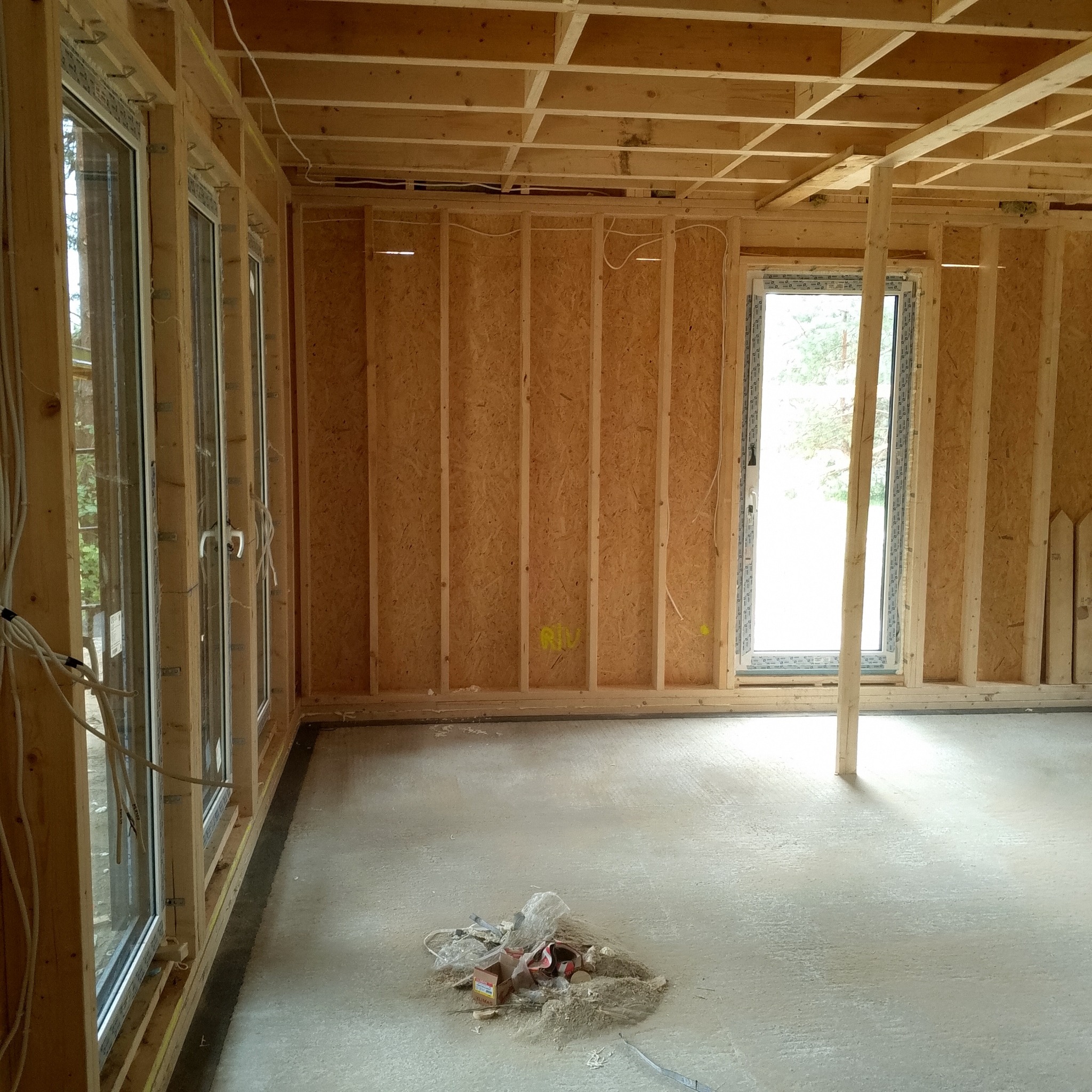 Wnętrze w stanie surowym: drewniana konstrukcja ścian z płyt OSB, okna PCV z widokiem na zieleń, betonowa podłoga z resztkami materiałów budowlanych, drewniany strop.