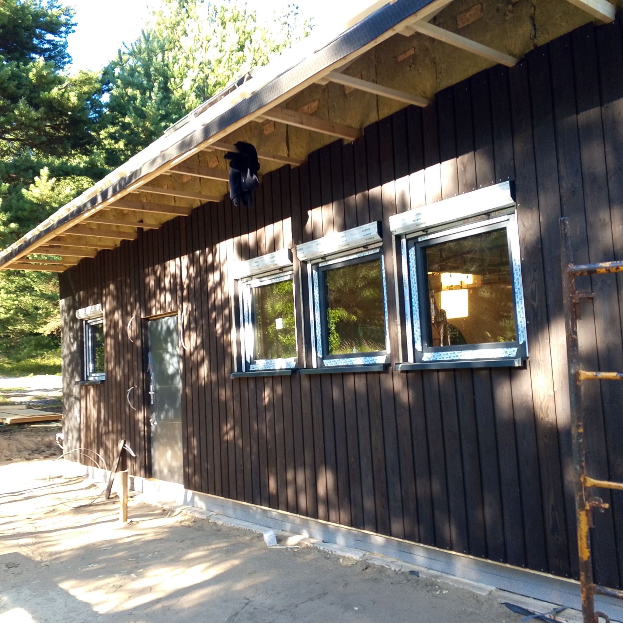 Budynek z ciemnego drewna z oknami i drzwiami, częściowo ocieplony, w trakcie budowy. Widoczne rusztowanie i materiały budowlane. Tło stanowią drzewa.