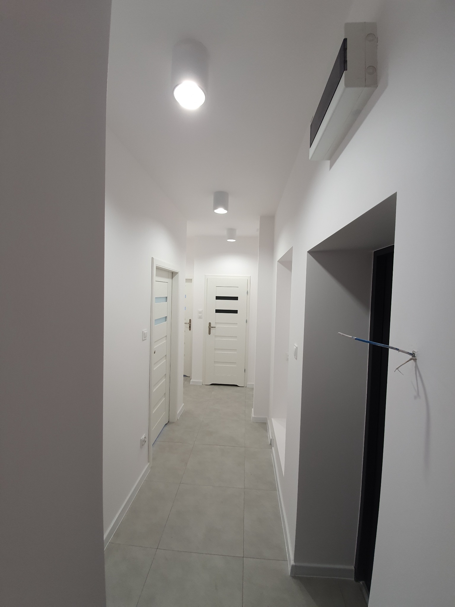Nowoczesny, długi korytarz z szarą podłogą z płytek, białymi ścianami i trzema białymi drzwiami, oświetlony okrągłymi lampami sufitowymi; widoczny fragment instalacji elektrycznej przy czarnej...