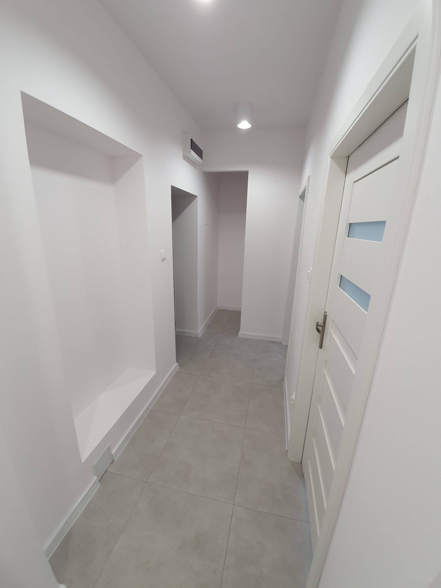 Wykończony korytarz w odcieniach bieli i szarości z widoczną wnęką w ścianie, szarą podłogą z płytek i białymi drzwiami z przeszkleniami.