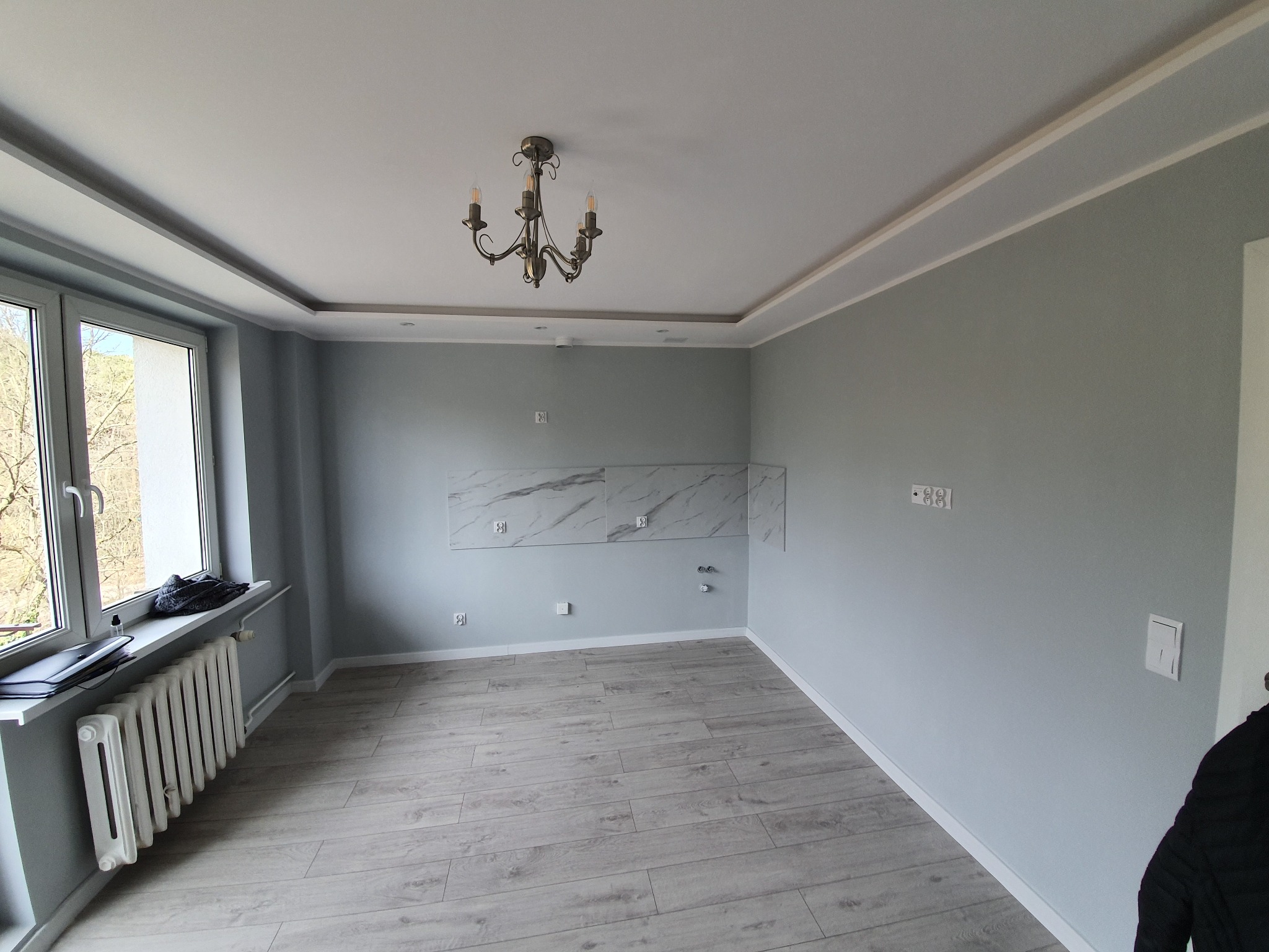 Wnętrze pokoju po remoncie: szare ściany, panele podłogowe w odcieniach szarości, biały grzejnik pod oknem, dekoracyjny żyrandol i marmurowe płytki na ścianie w miejscu planowanej kuchni.