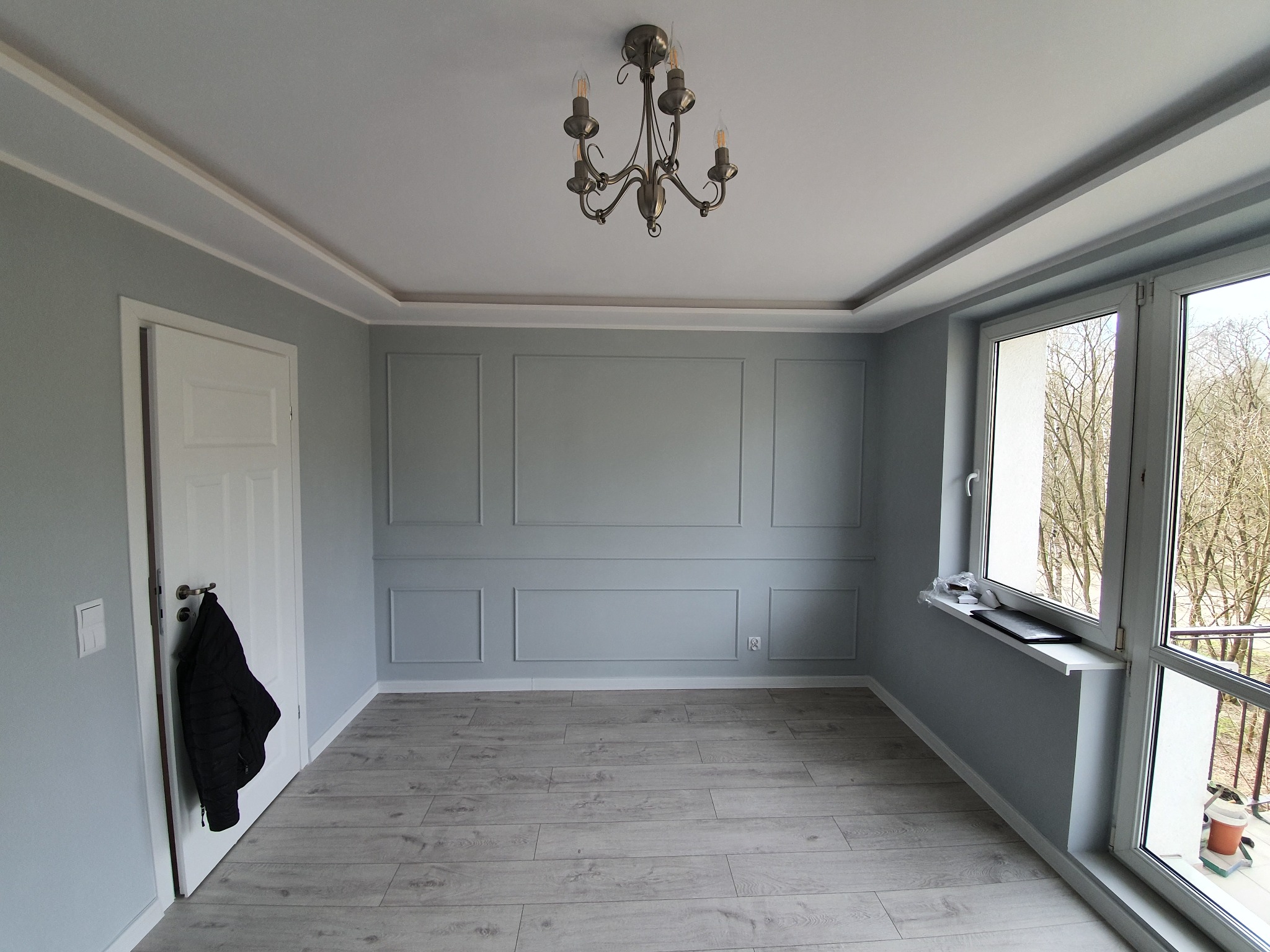 Jasnoszary pokój z panelami ściennymi, drewnianą podłogą, białymi listwami przypodłogowymi i sufitowymi, oraz żyrandolem w stylu retro. Na drzwiach wisi czarna kurtka, a na parapecie leżą przedmioty.