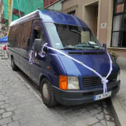 Niebieski bus udekorowany białymi wstążkami i kwiatami, zaparkowany na brukowanej ulicy w centrum miasta, prawdopodobnie do ślubu.