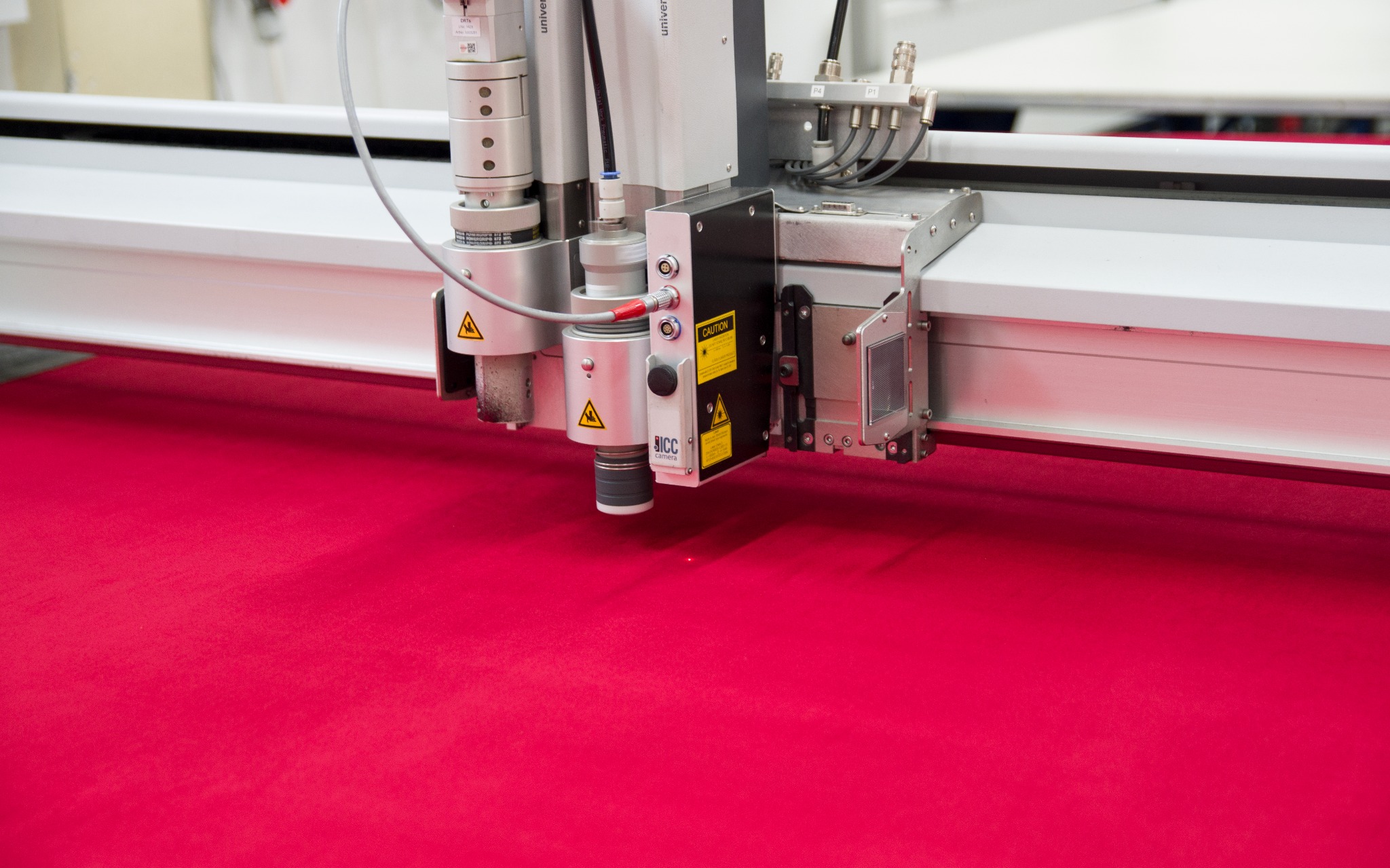 Automatyczna maszyna tnąca laserem precyzyjnie wycina wzór w intensywnie czerwonej tkaninie; widoczna głowica lasera z ostrzeżeniami i logotypem producenta.