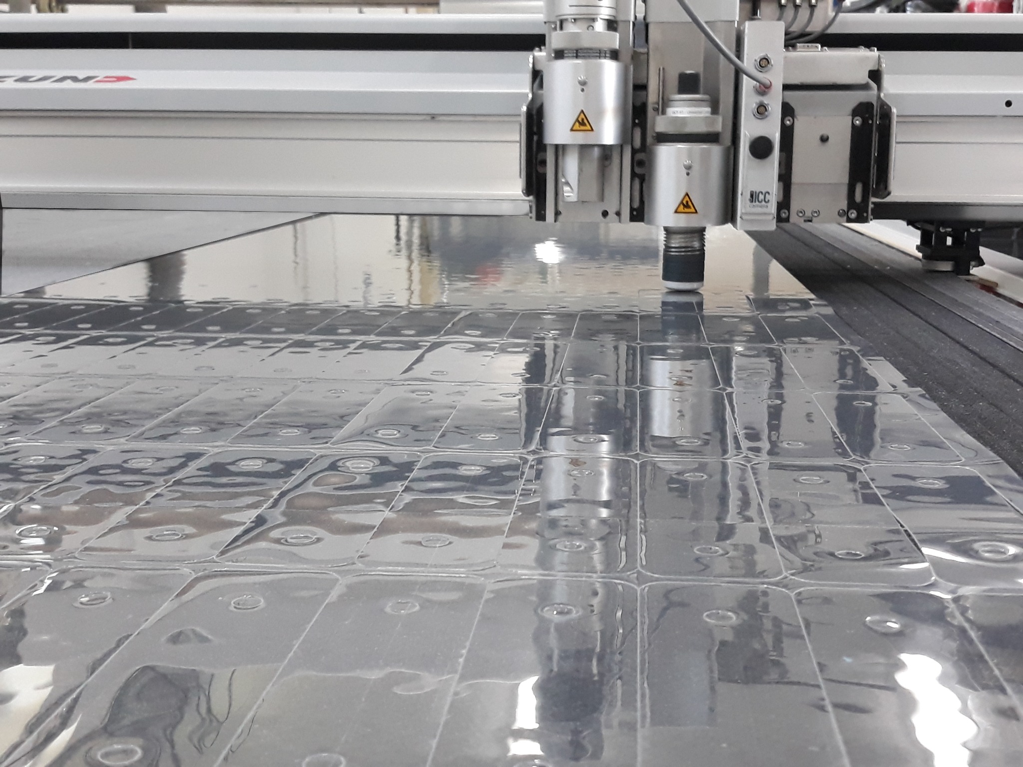 Precyzyjna maszyna tnąca ZUND wycina powtarzalny wzór w arkuszu transparentnego tworzywa sztucznego, widoczne liczne wgłębienia i refleksy świetlne na powierzchni materiału.