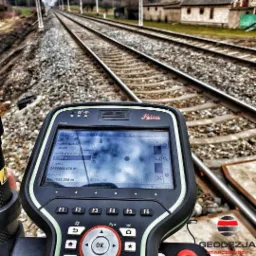 Kontroler geodezyjny Leica z widocznym ekranem pomiarowym na tle torów kolejowych i budynku w oddali. Widoczny logotyp firmy geodezyjnej w prawym dolnym rogu.