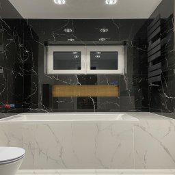 REMONO Usługi Remontowo-Wykonczeniowe - Nowoczesna łazienka z czarnymi marmurowymi płytkami, białą wanną i grzejnikiem drabinkowym. Złota mozaika nad wanną, oświetlenie punktowe na suficie.