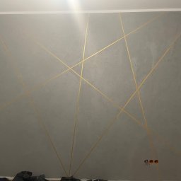 REMONO Usługi Remontowo-Wykonczeniowe - Szara ściana z dekoracyjnymi złotymi liniami tworzącymi geometryczny wzór, widoczne gniazdka elektryczne i fragment niezakończonego wykończenia przy drzwiach.