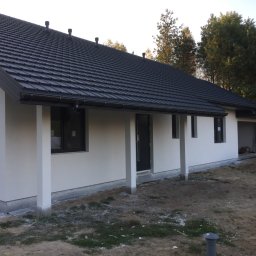 REMONO Usługi Remontowo-Wykonczeniowe - Nowoczesny parterowy dom z białymi ścianami i grafitowym dachem z blachodachówki, widok z zewnątrz na elewację frontową, teren wokół domu częściowo uporządkowany.