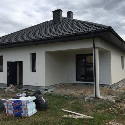 REMONO Usługi Remontowo-Wykonczeniowe - Parterowy dom jednorodzinny z szarą dachówką i świeżo otynkowaną białą elewacją, widoczne materiały izolacyjne i worki na gruz przed wejściem, sugerujące prace wykończeniowe.