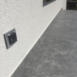 REMONO Usługi Remontowo-Wykonczeniowe - Zewnętrzne gniazdko elektryczne z szarą ramką na elewacji o strukturze baranka, z szarą, kamienną podłogą tarasu i czarną półką w tle.
