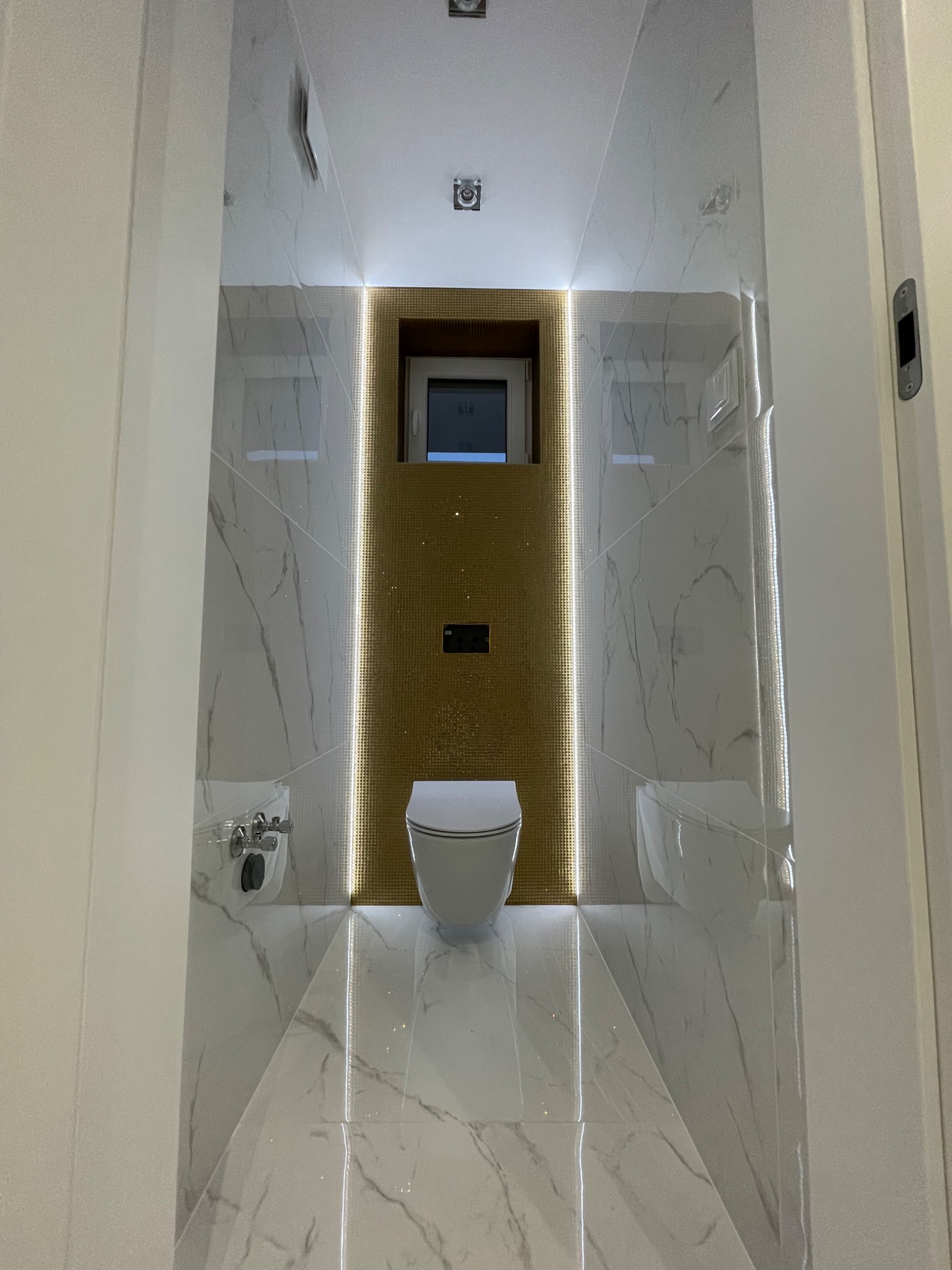 Nowoczesna łazienka w odcieniach bieli i złota z marmurowymi płytkami, podświetleniem LED wzdłuż ściany z mozaiką i wiszącą toaletą.
