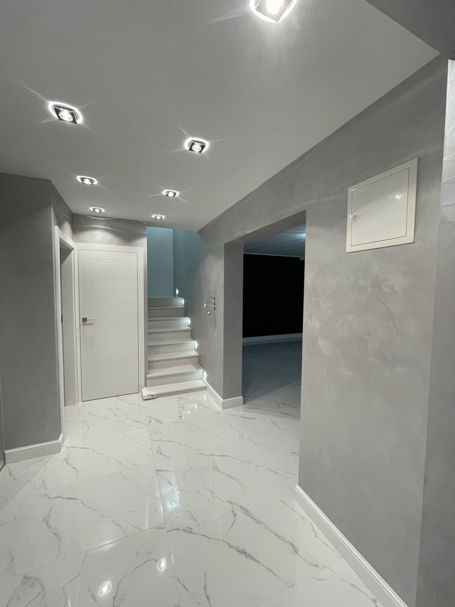 Nowoczesne wnętrze z marmurową podłogą, szarymi ścianami o dekoracyjnej fakturze, białymi drzwiami i podświetlanymi schodami prowadzącymi na wyższą kondygnację.