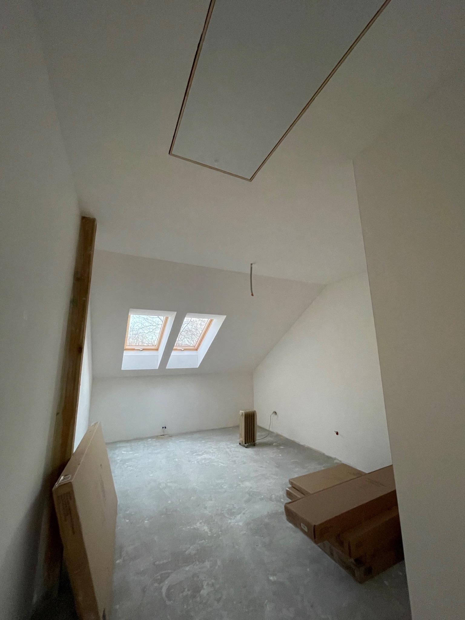 Poddasze w trakcie wykańczania: widoczne dwa okna dachowe, surowe ściany, niezagruntowana podłoga, skrzynki z materiałami budowlanymi oraz otwór włazowy w suficie.