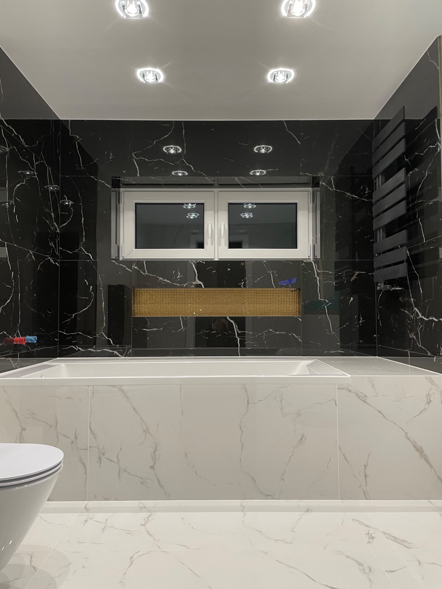 Nowoczesna łazienka z czarnymi marmurowymi płytkami, białą wanną i grzejnikiem drabinkowym. Złota mozaika nad wanną, oświetlenie punktowe na suficie.