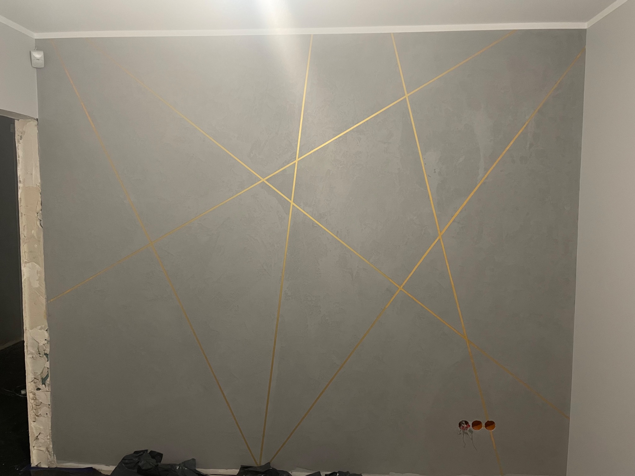 Szara ściana z dekoracyjnymi złotymi liniami tworzącymi geometryczny wzór, widoczne gniazdka elektryczne i fragment niezakończonego wykończenia przy drzwiach.