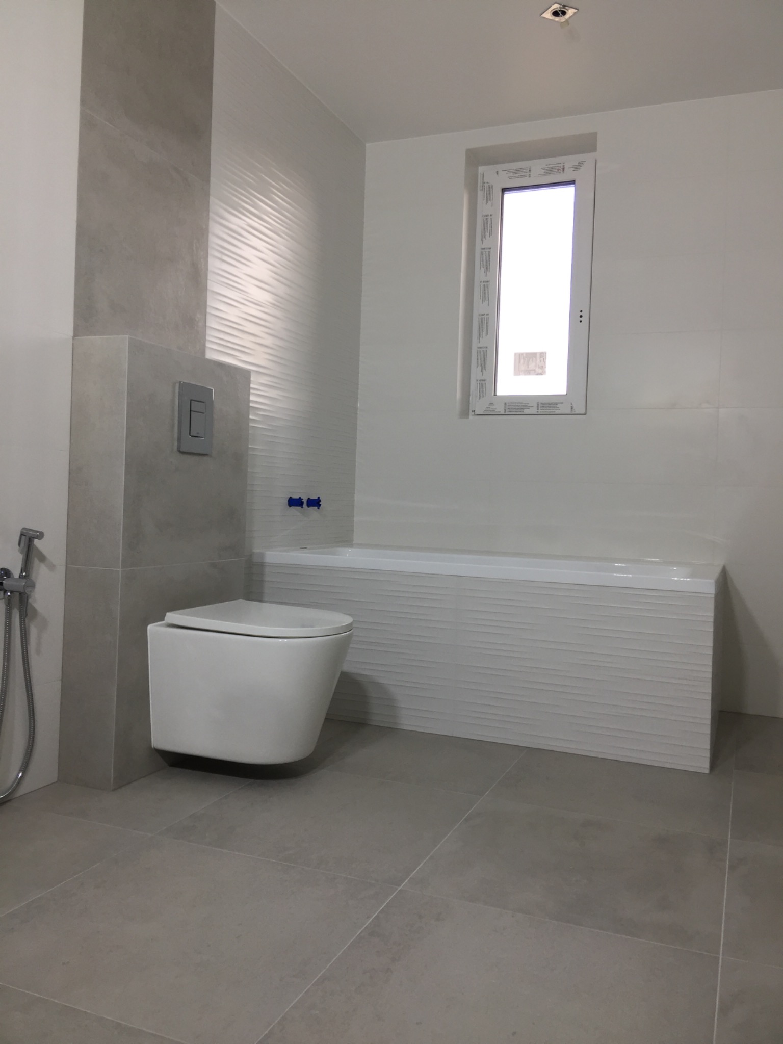 Nowoczesna, minimalistyczna łazienka z podwieszaną toaletą, wanną obudowaną płytkami z delikatnym wzorem fal oraz dużym oknem w białej ramie.