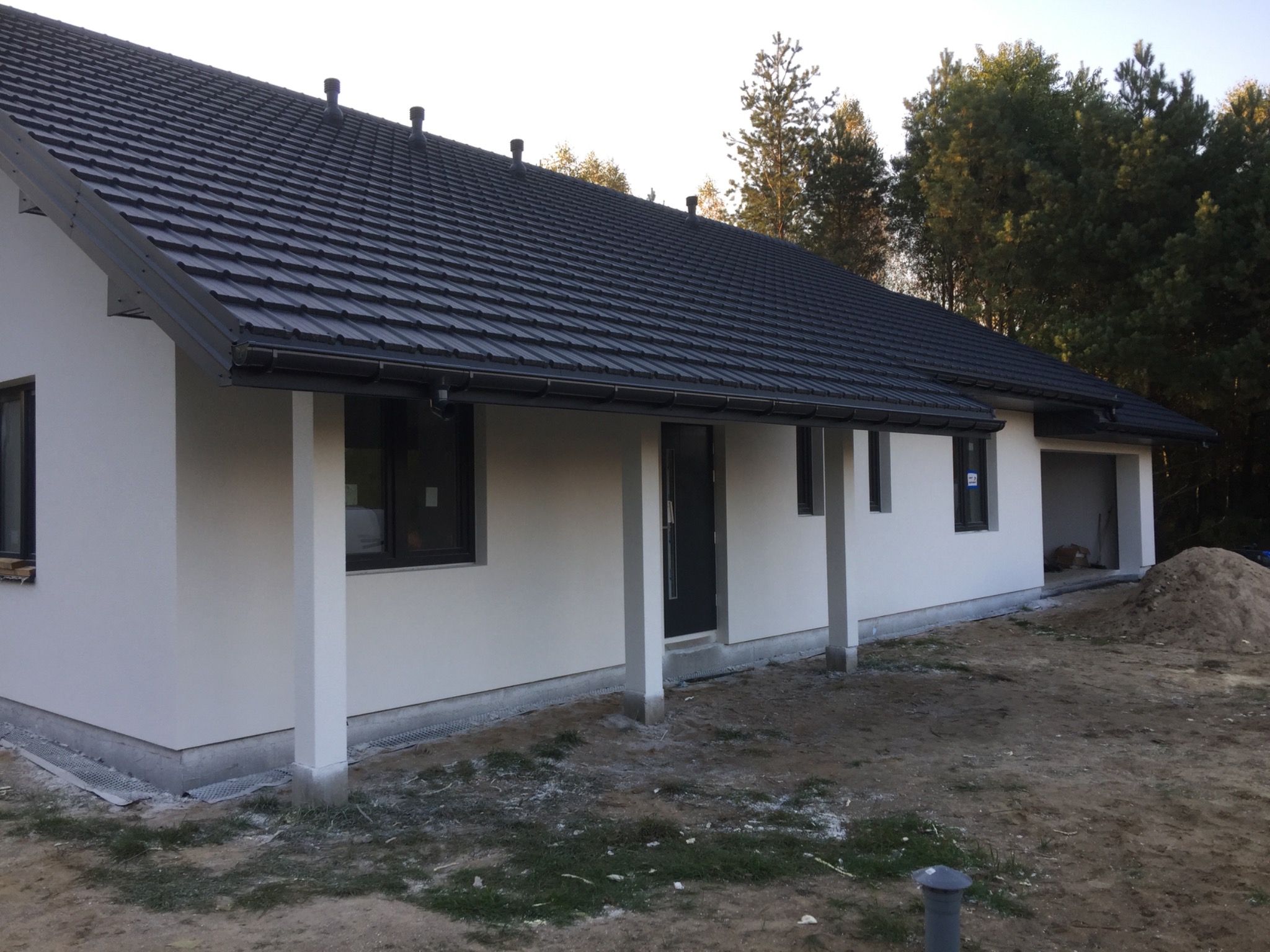 Nowoczesny parterowy dom z białymi ścianami i grafitowym dachem z blachodachówki, widok z zewnątrz na elewację frontową, teren wokół domu częściowo uporządkowany.