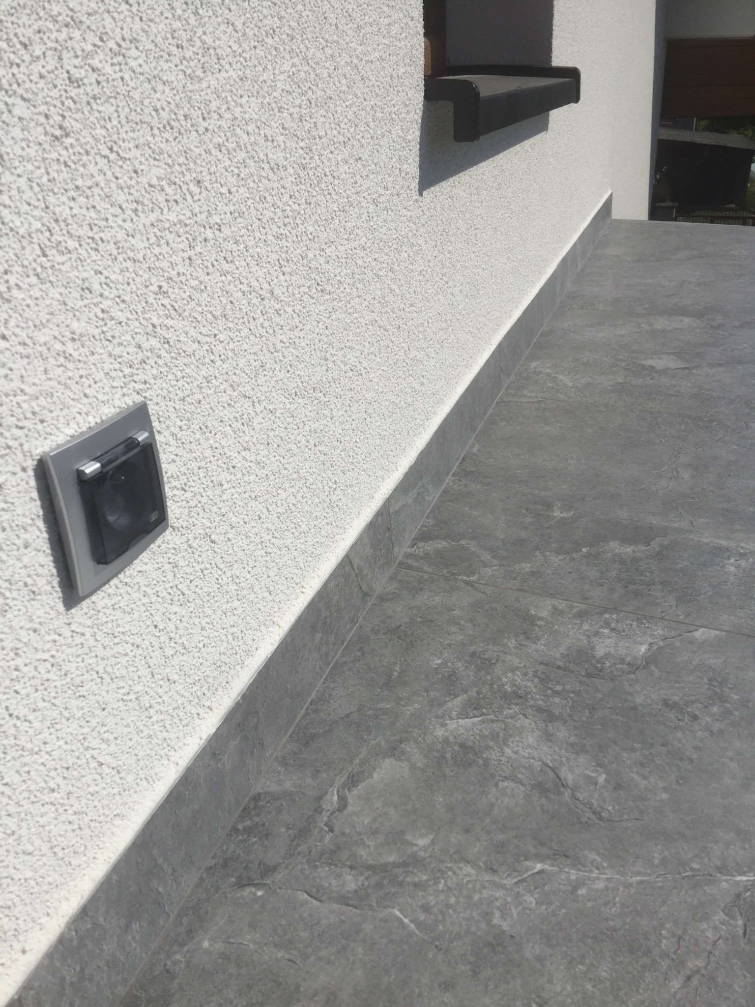 Zewnętrzne gniazdko elektryczne z szarą ramką na elewacji o strukturze baranka, z szarą, kamienną podłogą tarasu i czarną półką w tle.