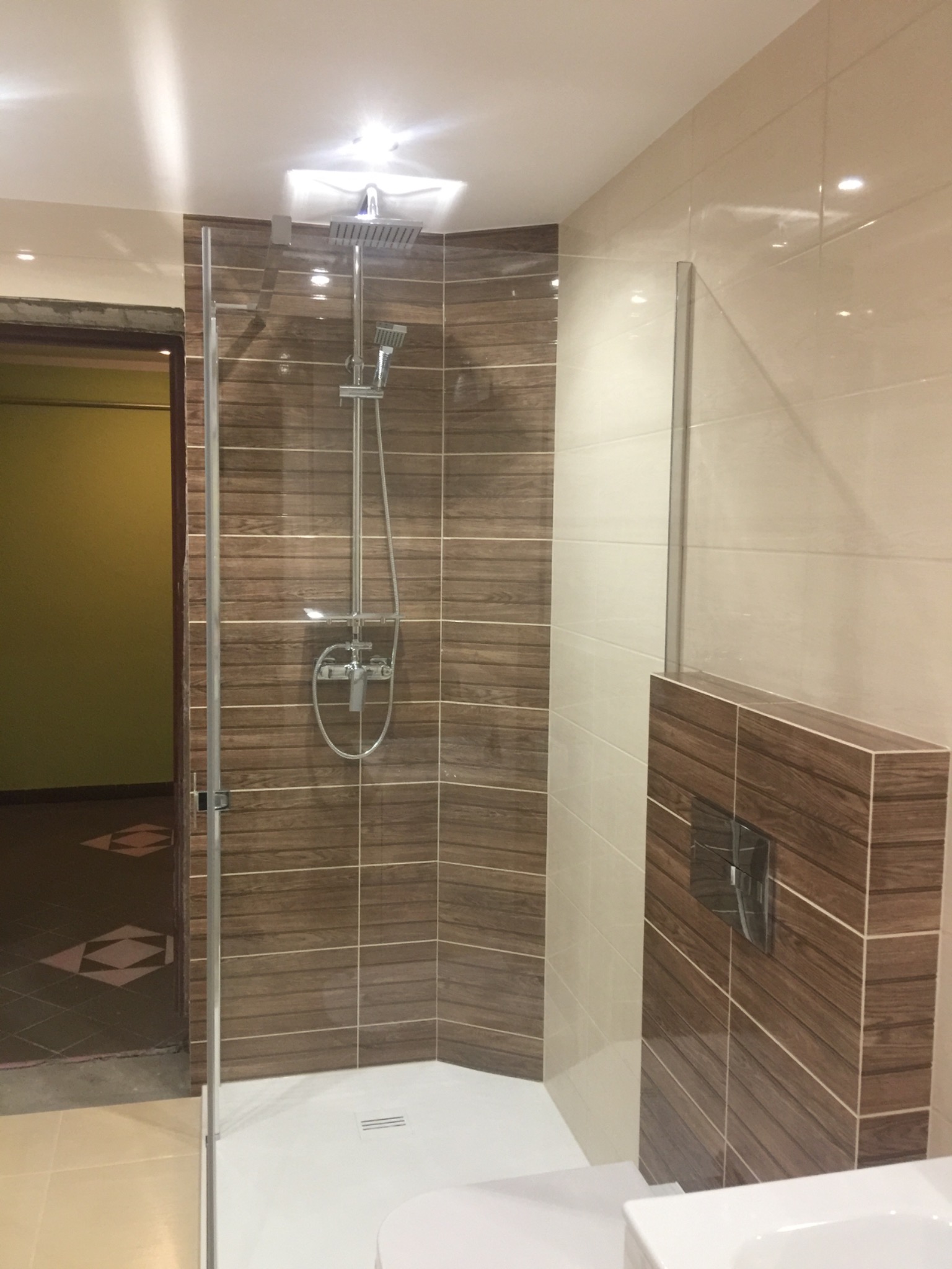 Nowoczesna kabina prysznicowa z deszczownicą i panelami imitującymi drewno, widok na fragment łazienki z zieloną ścianą i wzorzystą podłogą.