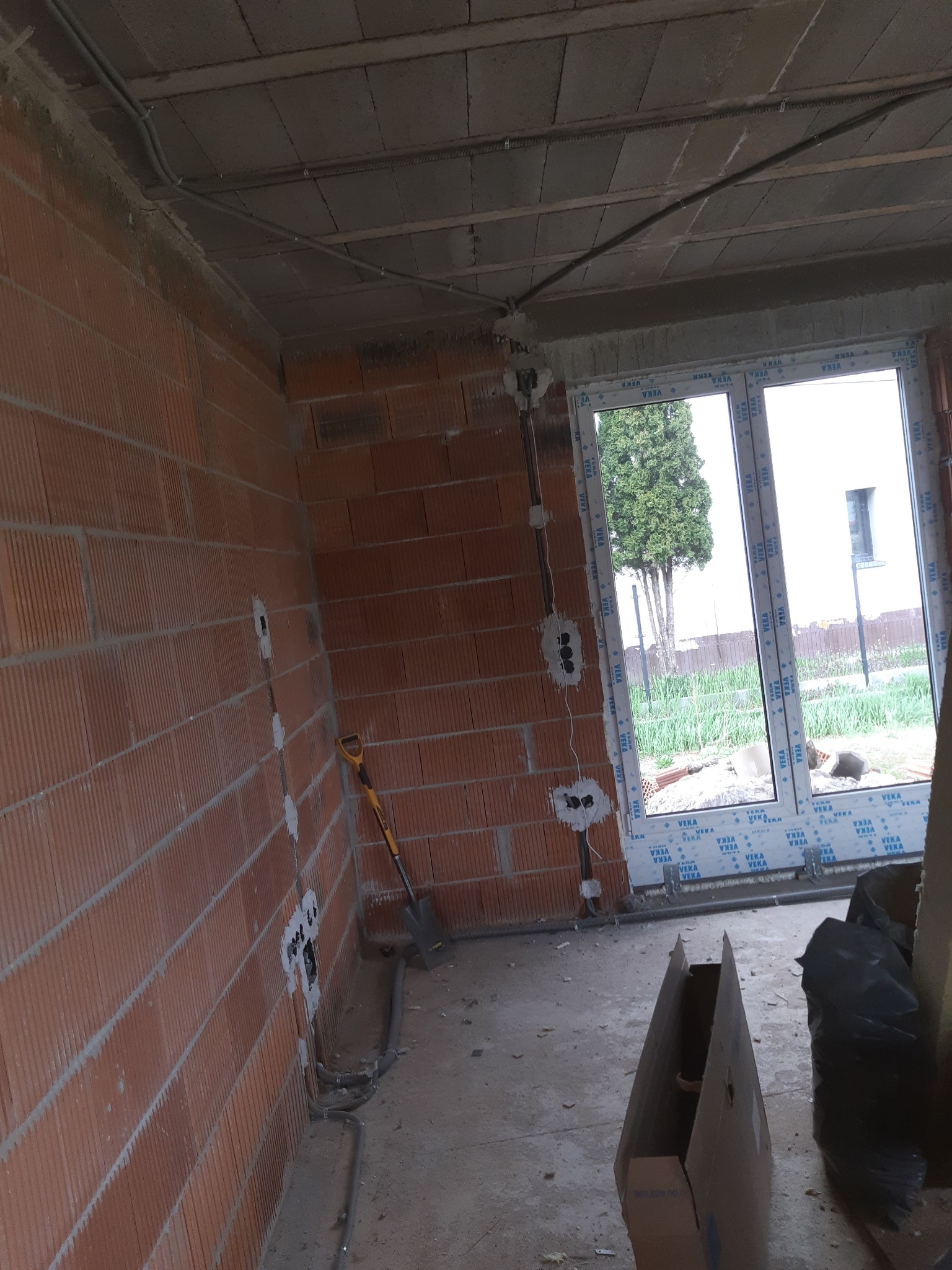Wnętrze w trakcie prac wykończeniowych, widoczne przewody elektryczne prowadzone po ścianie z cegły i pod sufitem, puszki instalacyjne w ścianie, szpadel oparty o ścianę, okno PCV z widokiem...