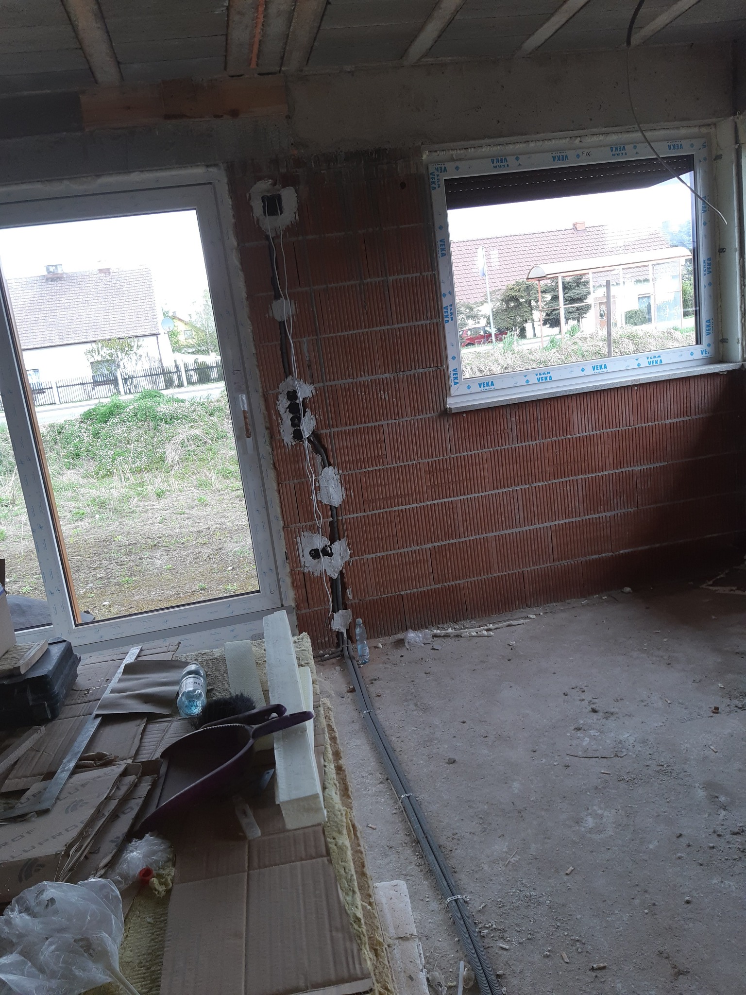 Wewnętrzne okablowanie elektryczne w surowym budynku z widocznymi puszkami instalacyjnymi i przewodami, okno z szybą zespoloną i ceglaną ścianą w tle.