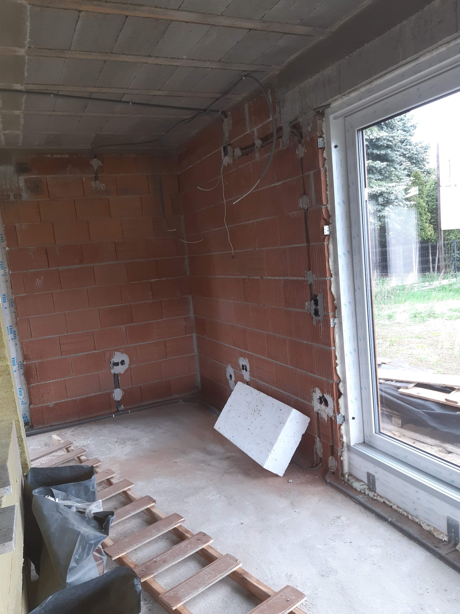 Wnętrze w trakcie prac instalacyjnych: widoczne przewody elektryczne w ścianach z czerwonej cegły, okno PCV, fragment rusztowania i styropian na posadzce.