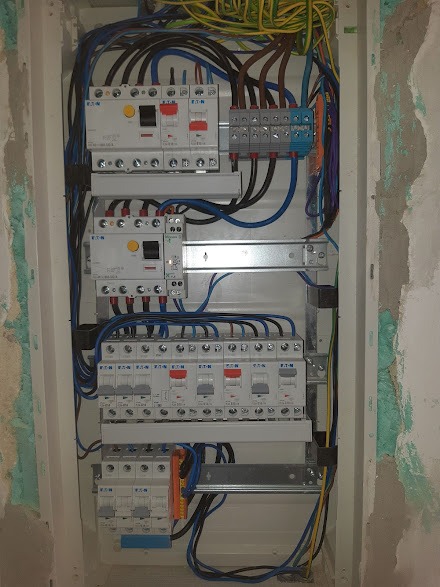 Otwarta skrzynka elektryczna z zamontowanymi bezpiecznikami EATON, przewodami w kolorach niebieskim, czarnym i żółto-zielonym, widoczna na tle surowej ściany w trakcie wykończenia.